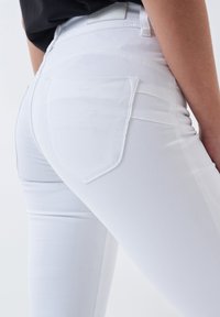 Jean blanc avec un design ajusté, une texture lisse et des poches arrière avec des coutures contrastantes. Le matériau semble extensible et moulant.