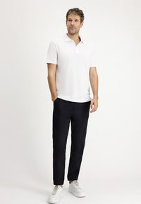 Biała koszulka polo z krótkim rękawem i kołnierzykiem, połączona z granatowymi spodniami slim fit i białymi trampkami, stoi na neutralnym tle.