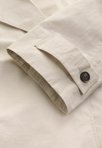 Beige bomuldsstof med en foldet manchet, prydet med en mørk knap og kontrastsyninger langs kanterne. Glat tekstur, minimalistisk design.