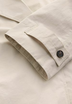 Beige katoenen stof met een omgevouwen manchette, voorzien van een donkere knoop en contrasterende stiksels langs de randen. Gladde textuur, minimalistisch ontwerp.
