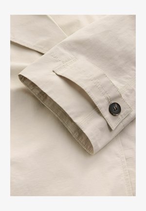 Tissu en coton beige avec un poignet plié, comportant un bouton foncé et des coutures contrastées le long des bords. Texture lisse, design minimaliste.