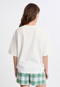 Donna con lunghi capelli castani indossa una maglietta bianca larga e pantaloni corte a scacchi verdi e bianchi, in piedi contro uno sfondo bianco semplice, con la schiena rivolta verso la macchina fotografica.