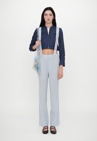 Jaqueta de ganga escura cropped com costura em forma de coração, calças cinza claro de corte elegante e uma mala transparente com desenhos coloridos e detalhes de alças.