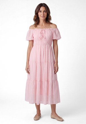 Woman wearing a light pink off-shoulder midi dress with puffed sleeves and beige flats, standing against a plain white background.