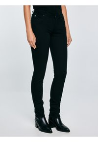 Mujer con jeans negros ajustados y botines negros con tacón, de pie contra un fondo claro y liso.