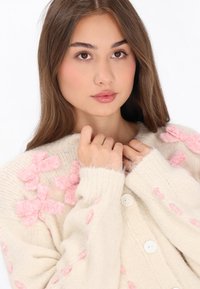 Beige gebreide cardigan met textuur in de vorm van roze strikapplicaties, een knoopsluiting en geribde manchetten. Zachte stof met een gezellige uitstraling.