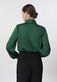 Femme aux cheveux châtains attachés en chignon, portant un chemisier en satin vert foncé à manches longues et un pantalon noir taille haute, vue de dos sur un fond blanc.