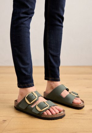 Fødder iført mørkegrønne sandaler med dobbelte stropper og store guldbeslag, kombineret med mørkeblå skinny jeans, stående på trægulv.