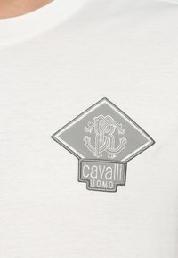 Biała bawełniana koszulka z szarym, haftowanym logo w kształcie diamentu o skomplikowanym wzorze oraz napisem "cavalli UOMO" na dole.