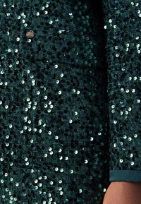 Mörkgrön paljettklädd tyg med en texturerad yta, prydd med glänsande runda paljetter i varierande nyanser. En liten metalllogotyp kan ses.