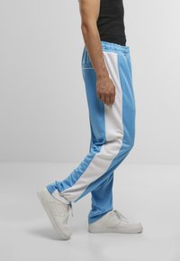 Hellblaue Jogginghose mit weißen Seitenstreifen, aus glattem Stoff gefertigt. Sie hat einen elastischen Bund und eine bequeme Passform, kombiniert mit weißen Turnschuhen.
