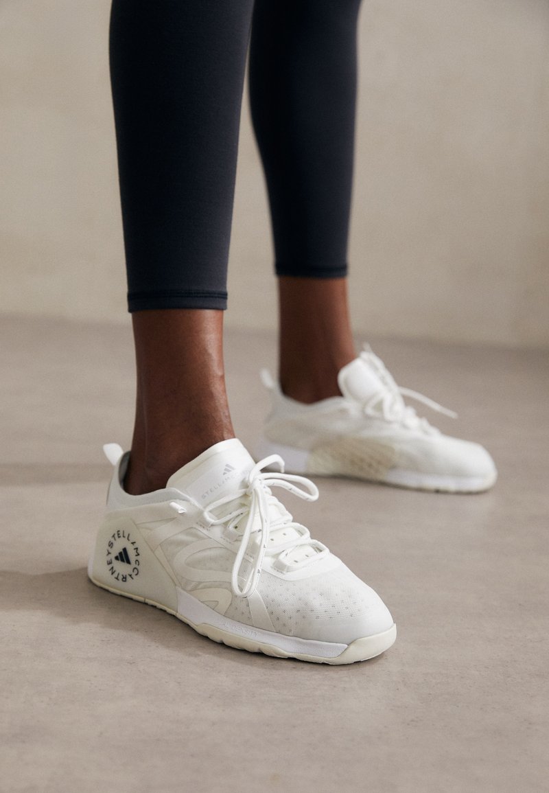 Fekete, testhez simuló leggingset viselő személy fehér fűzős sportcipőben, fekete logókkal, áll egy világosszürke padlón.