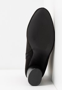 Botín negro de ante hasta el tobillo con una suela de goma negra texturizada y un tacón bajo y redondo. Cuenta con un panel lateral elástico para facilitar el uso.