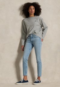 Mujer con cabello rizado que lleva una camiseta gris de manga larga, vaqueros azul claro y zapatillas azul marino, de pie contra una pared neutra.