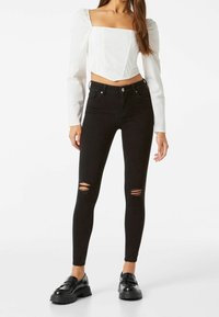 Jeans negros ajustados con detalles desgastados, combinados con una blusa corta blanca con escote cuadrado y mangas abullonadas. Textura suave.
