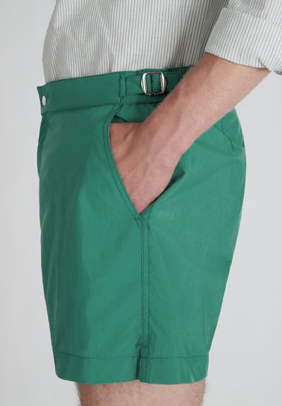 Hombre que lleva puestos unos pantalones cortos verdes con detalle de hebilla lateral, con la mano en el bolsillo, combinado con una camisa de rayas azul claro metida por dentro.