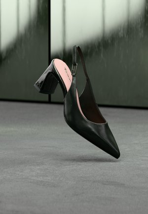 Black patent leather slingback shoe with pointed toe and block heel, suspended above gray carpet against a greenish reflective background.