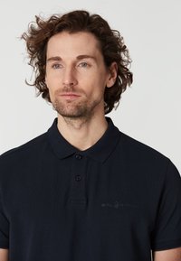 Marinblå polo med knäppbar krage, två knappar och en diskret broderad 'SAIL RACING'-logo på vänster bröst.