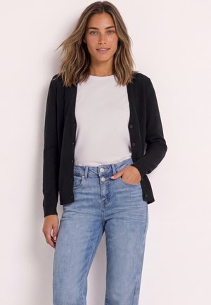 Femme aux cheveux mi-longs portant une chemise blanche, un cardigan noir boutonné et un jean taille haute bleu clair, une main dans la poche.