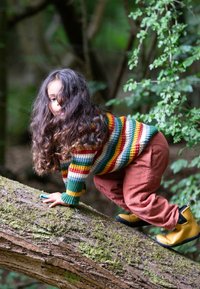 Kind in einem bunt gestreiften Strickpullover, terrakottafarbenen Hosen und gelben Gummistiefeln, das auf einem von Moos bedeckten Stamm klettert, umgeben von Grün.