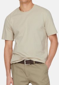 Beige Baumwoll-T-Shirt mit kurzen Ärmeln und Rundhalsausschnitt, kombiniert mit khakifarbenen Hosen und einem braunen Gürtel mit einer silbernen Schnalle.
