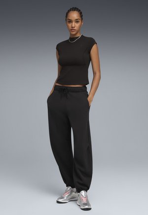 Mujer de pie con las manos en los bolsillos, vistiendo una camiseta sin mangas negra, pantalones deportivos negros y zapatillas plateadas con cordones rosas sobre fondo gris.