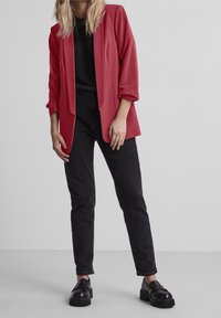 Červený blazer s ohrnutými rukávy, černý top a černé džíny. Obuv je ve stylu robustních, lesklých černých mokasín. Jednoduchý, střihově přesný design.