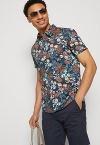 Korte ærmer, skjorte med knaplukning, der har et blomstermotiv i nuancer af blåt, rødt og hvidt mod en mørk marineblå baggrund, lavet af let materiale.