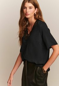 Blouse noire à manches courtes et décolleté en V, dotée d'une bordure en dentelle le long de l'encolure. Tissu lisse, associée à une jupe en cuir.