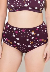 Frau in hochtailliertem lila Unterwäsche und passendem BH mit weißen, rosa und senffarbenen Polka-Dots vor einem einfarbigen Hintergrund.