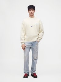 Jeune homme portant un pull écru avec un texte bleu clair, un jean bleu clair usé et des chaussures marron foncé avec des lacets rouges, debout avec une main dans la poche.