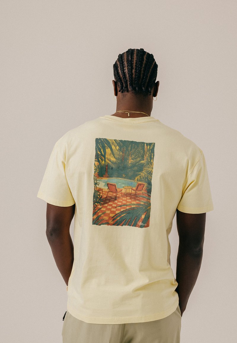 Homme aux cheveux tressés porte un t-shirt jaune pâle avec un imprimé patio tropical avec deux chaises et une table près d'une piscine.