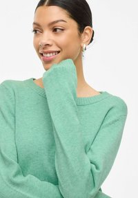 Mujer sonriente con cabello oscuro, con un suéter verde, descansando suavemente la barbilla en la mano, contra un fondo blanco liso.