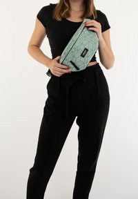 Marsupio green con motivo geometrico e zip, tenuto in mano. Presenta un'etichetta del marchio nera; indossato con pantaloni neri a vita alta.