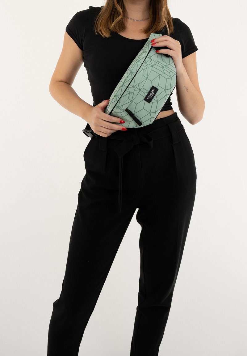 Marsupio green con motivo geometrico e zip, tenuto in mano. Presenta un'etichetta del marchio nera; indossato con pantaloni neri a vita alta.
