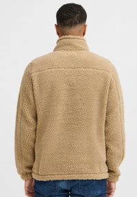 Beige fleece jacket with a high collar, textured surface, and relaxed fit. Features a hemmed bottom and long sleeves.