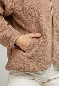 Veste en polaire couleur brun clair avec une surface texturée. Équipée de poches zippées et d'une bordure en zip beige lisse. Coupe standard.