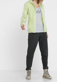 Giacca a zip verde chiaro con cappuccio, abbinata a una canotta grigia con grafica e pantaloni neri affusolati. Stivali da trekking color sabbia. Abbigliamento casual per attività all'aperto.