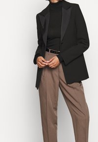 Frau in einem schwarzen Blazer mit satinierten Revers, schwarzem Rollkragenpullover und hochgeschnittenen braunen Hosen mit schwarzem Gürtel, die die Hände vorne gefaltet hat.