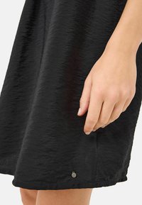 Svart, texturerad klänning med en hand som greppar fållen, med en liten metallaccent på sidan och en flödande, skrynklig tygutseende.