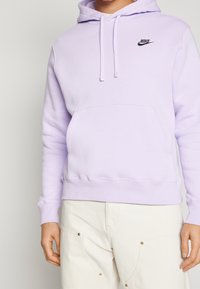 Person som bär en ljuslila Nike hoodie med framficka och krämfärgade byxor med nitar, visad från axlarna till mitten av låret.