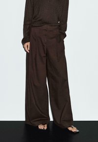 Wijde broek van donkerbruin stof, met plooien, hoge taille en een soepele textuur. Gdragen met minimalistische sandalen.