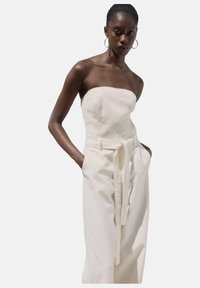 Combinaison sans bretelles en tissu crème clair avec une taille ceinturée, un design à jambe droite et des poches latérales. Texture lisse et silhouette élégante.