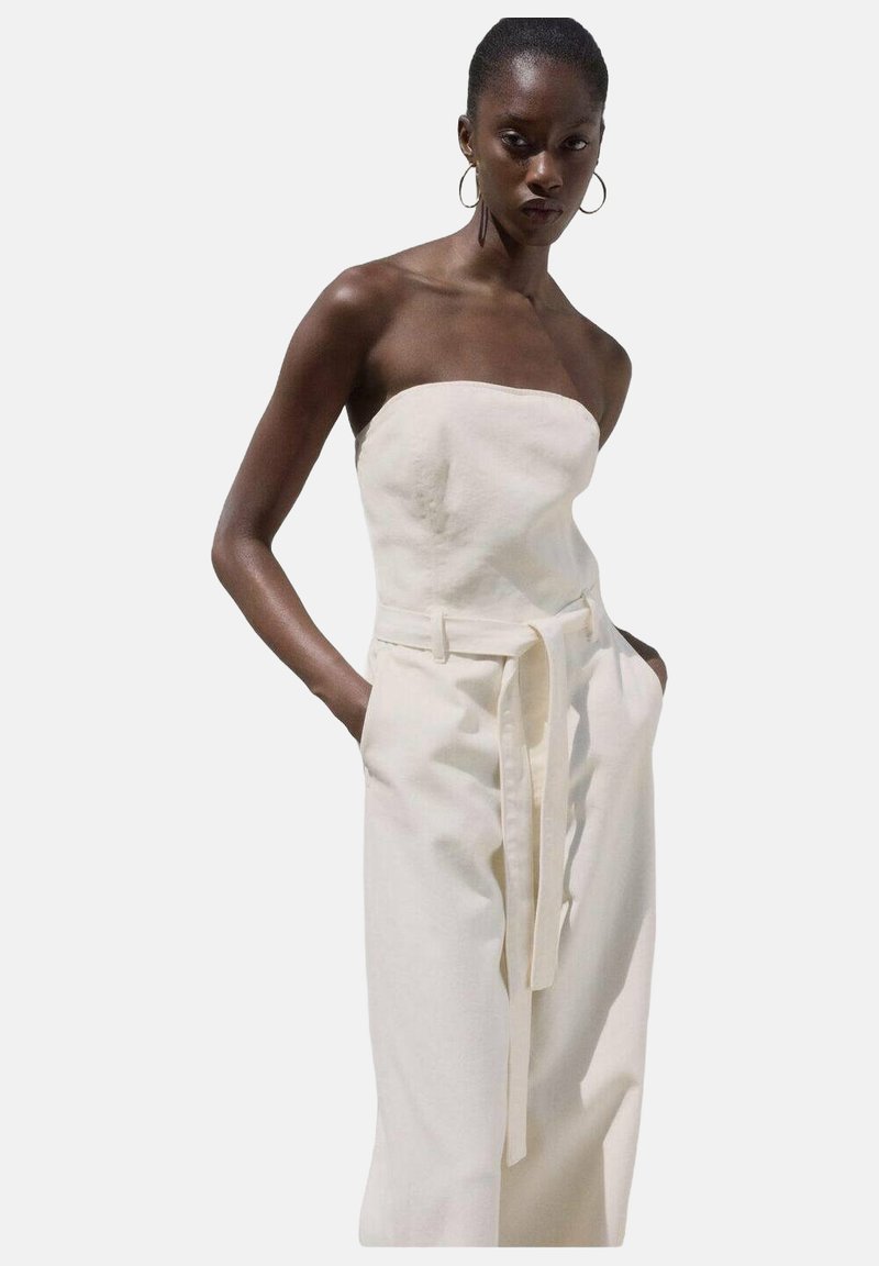 Combinaison sans bretelles en tissu crème clair avec une taille ceinturée, un design à jambe droite et des poches latérales. Texture lisse et silhouette élégante.
