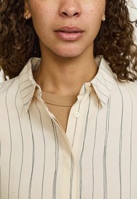 Cămașă bej cu dungi verticale negre, guler clasic și închidere cu nasturi. Modelul poartă un maieu bej deschis. Textură fină a țesăturii.