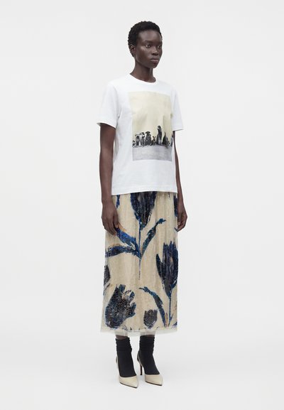 Femme portant un T-shirt blanc avec une photo de chien imprimée, une jupe beige avec de grands motifs floraux bleus, des chaussettes noires et des chaussures blanches à talons hauts.