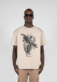 Beige bomulds t-shirt med et sort og gråt grafisk design af en engel med vinger og kyrillisk tekst, korte ærmer og en afslappet pasform.