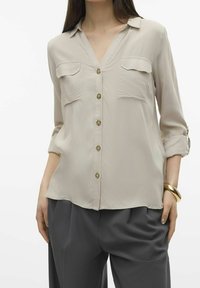 Blusa lunga beige chiaro con scollatura a V, due tasche sul petto, bottoni dorati e maniche arrotolate, abbinata a pantaloni grigi a gamba larga.