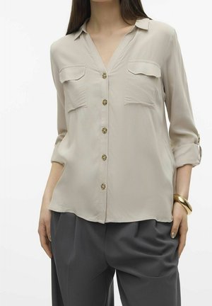 Blusa lunga beige chiaro con scollatura a V, due tasche sul petto, bottoni dorati e maniche arrotolate, abbinata a pantaloni grigi a gamba larga.