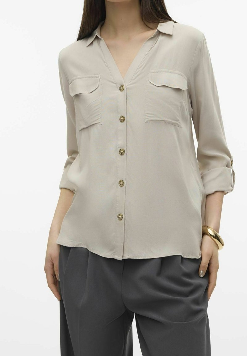 Blusa lunga beige chiaro con scollatura a V, due tasche sul petto, bottoni dorati e maniche arrotolate, abbinata a pantaloni grigi a gamba larga.